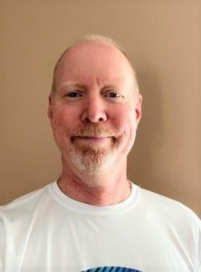 Man with a goatee smiling, wearing a white shirt against a beige background.