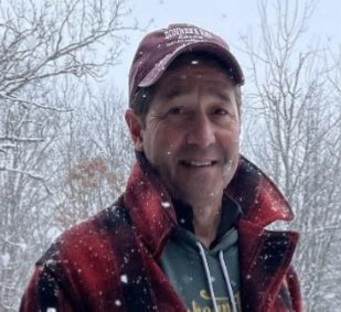 Man in plaid coat and cap smiles in a snowy outdoor setting.