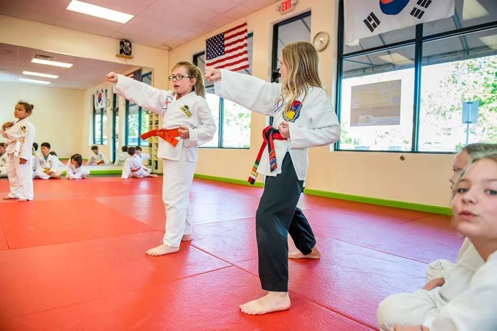 Children practicing martial arts in a dojo, one with a red belt, others watching.