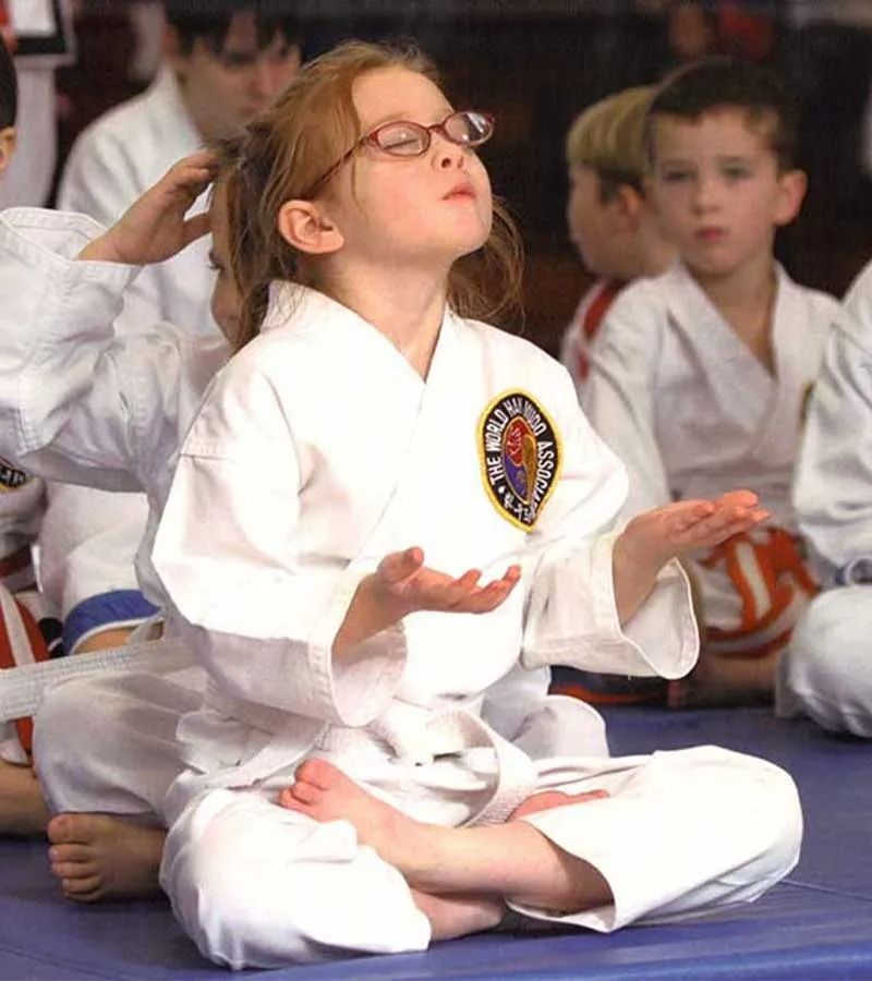 Girl in karate uniform meditating with eyes closed, hands up. Other children in background.