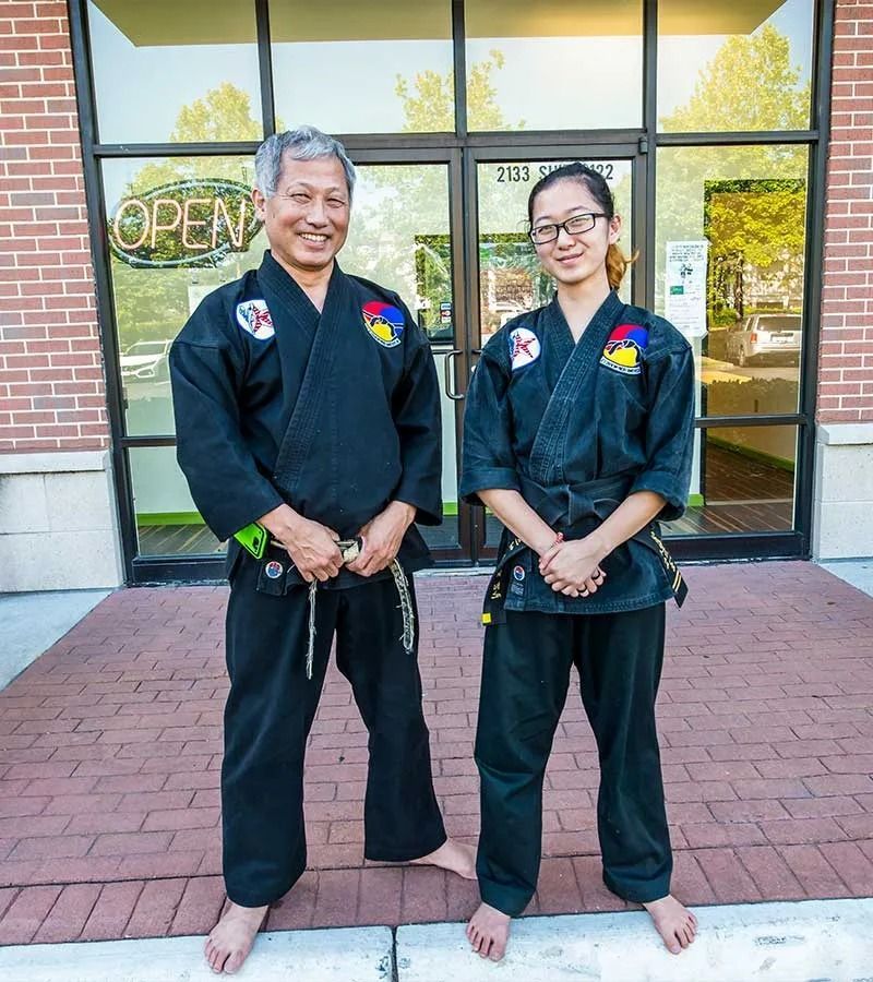 Two people in black martial arts uniforms stand outside a storefront; 