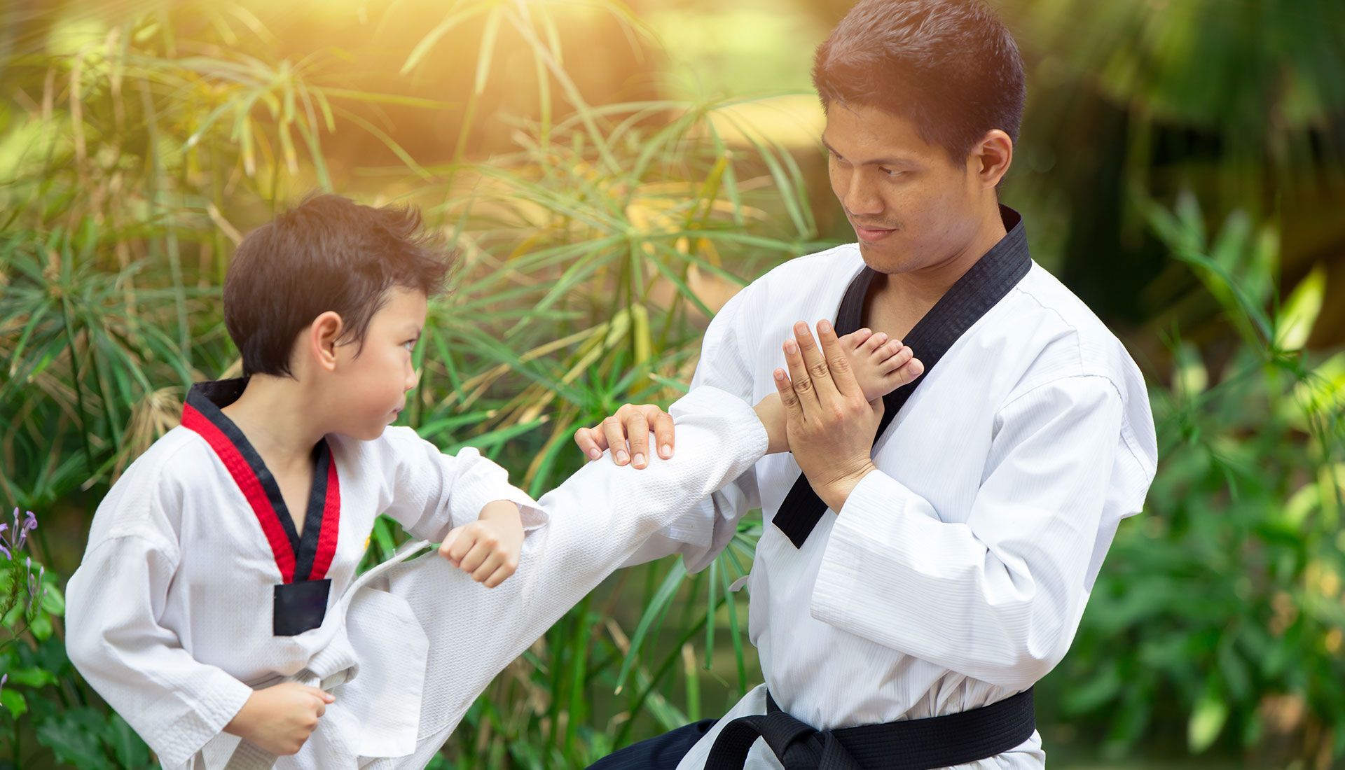 A person with a black belt teaches a child a Taekwondo kick outdoors.