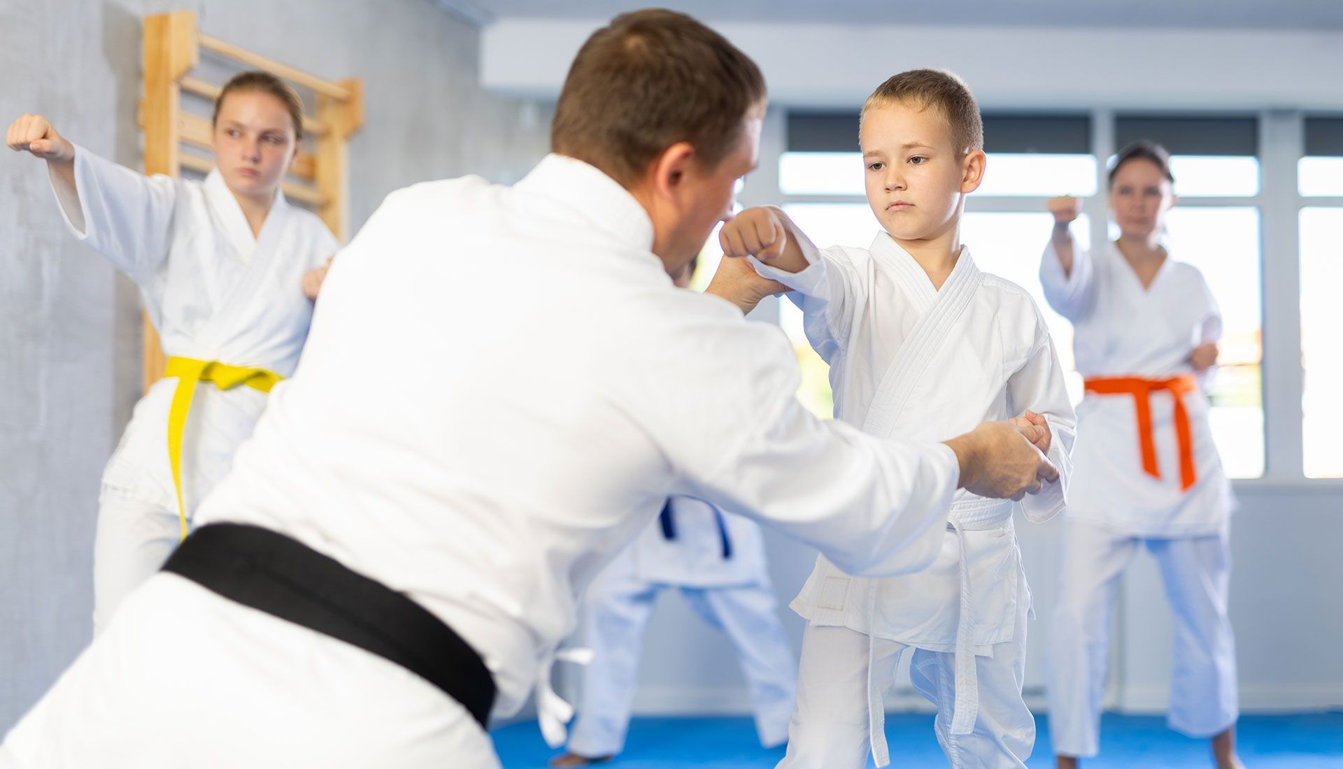 Martial arts class; instructor guides boy's punch, others practice in background. White gis, colorful belts, gym setting.