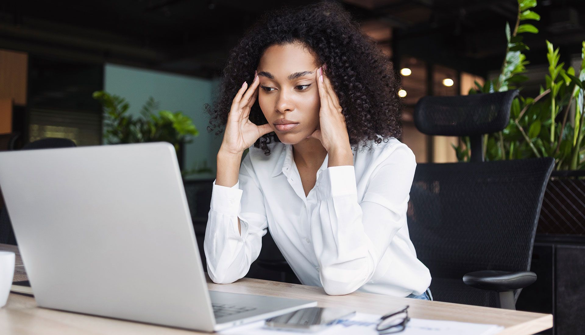 Woman with dark curly hair looking stressed, hands on her head, in front of a laptop at an office desk.