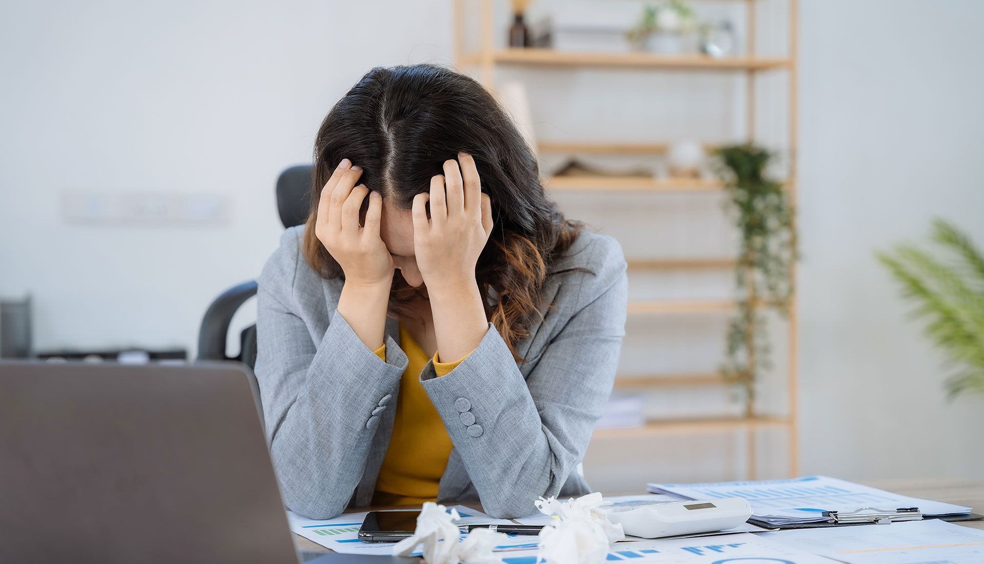 Woman in grey blazer at desk, hands on face, looking stressed.