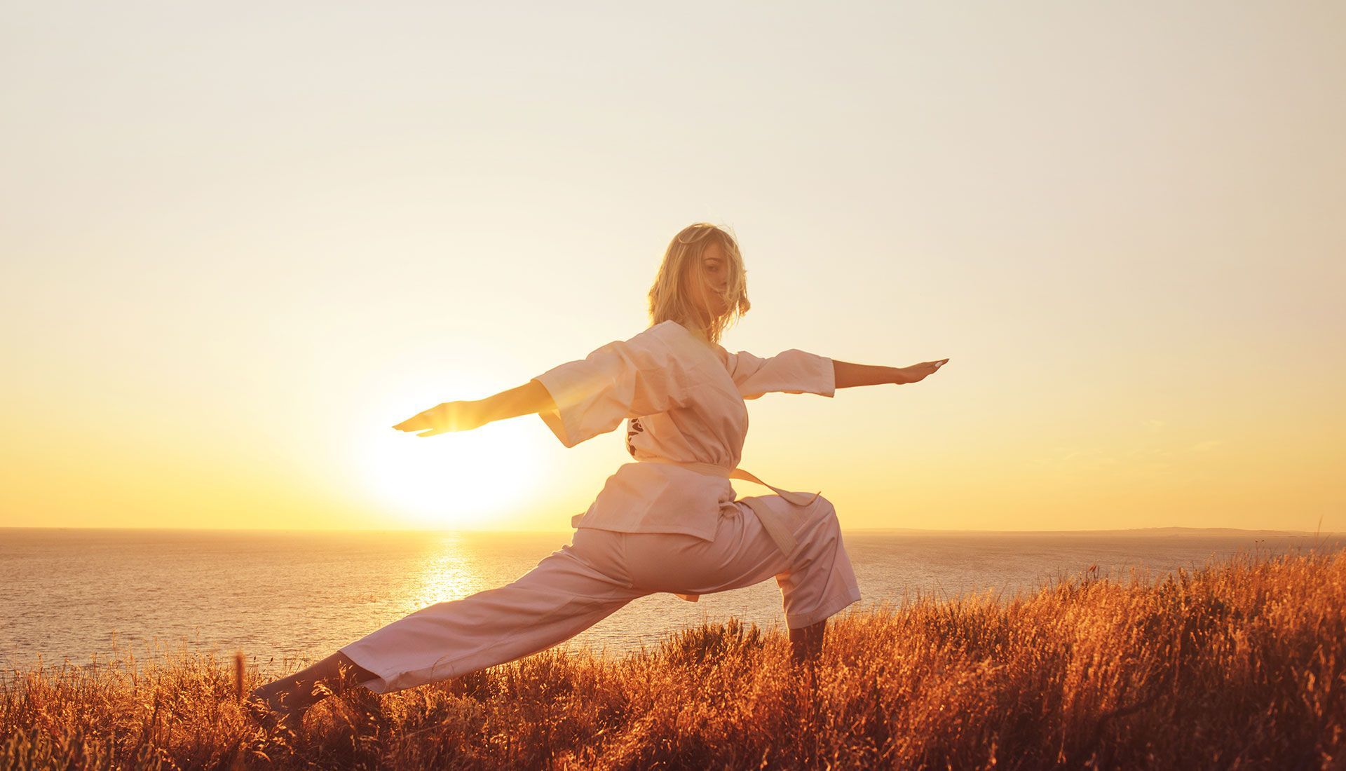 Woman in white karate gi in a low stance with arms outstretched, backlit by a sunset over the water.