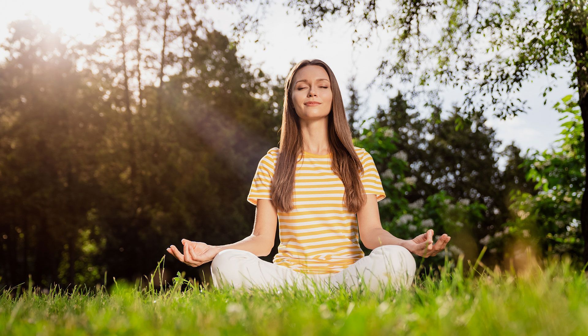 Woman meditating cross-legged on grass in a sunlit park, eyes closed, hands in a mudra position.