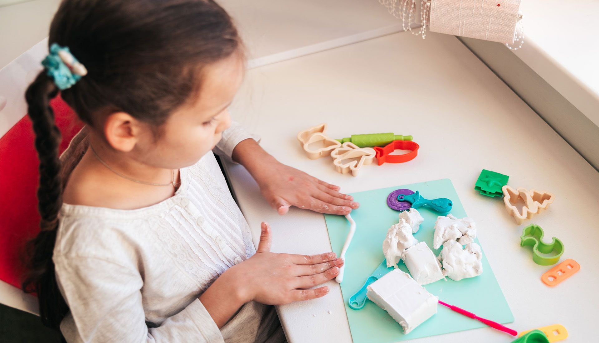 Girl seated at a table, molding clay on blue paper, with tools and colorful clay pieces nearby.