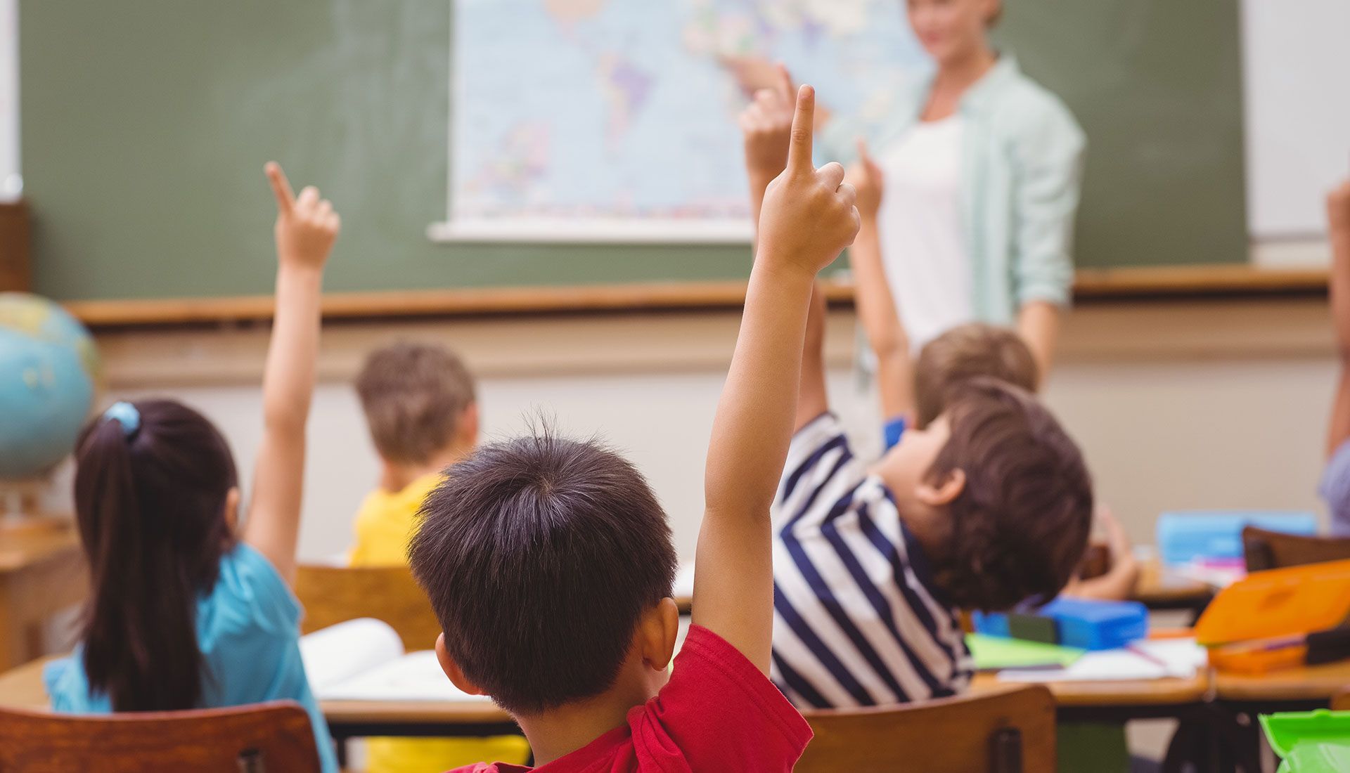 Students in a classroom raise their hands as a teacher points to a world map on a chalkboard.