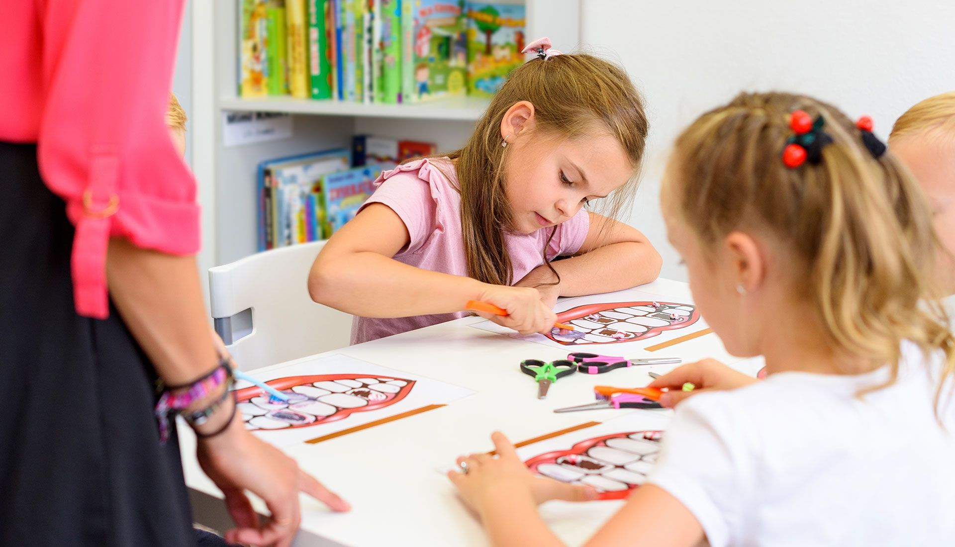 Children crafting at a white table in a classroom with a teacher's guidance.