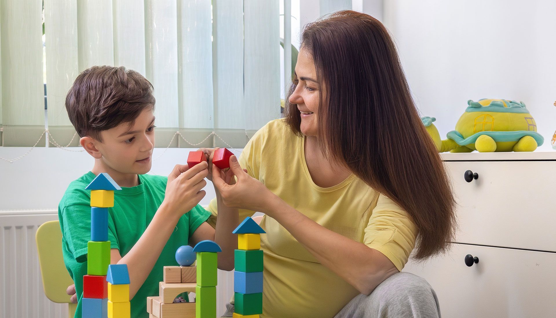 Woman and child building with colorful wooden blocks at a table. Child is focused; woman smiles.