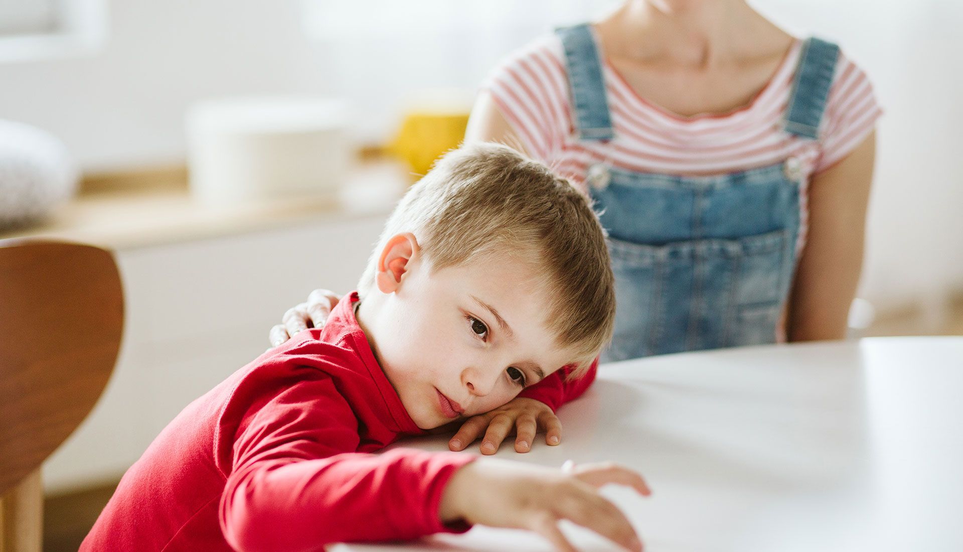 Boy in red shirt resting head on table, arm extended. Woman in denim overalls sits nearby.