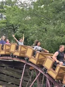 People on a wooden roller coaster, arms raised in excitement. Green trees in the background.