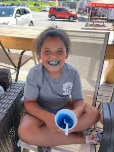 Girl smiles with blue lips, holding a cup of blue liquid, seated outdoors.