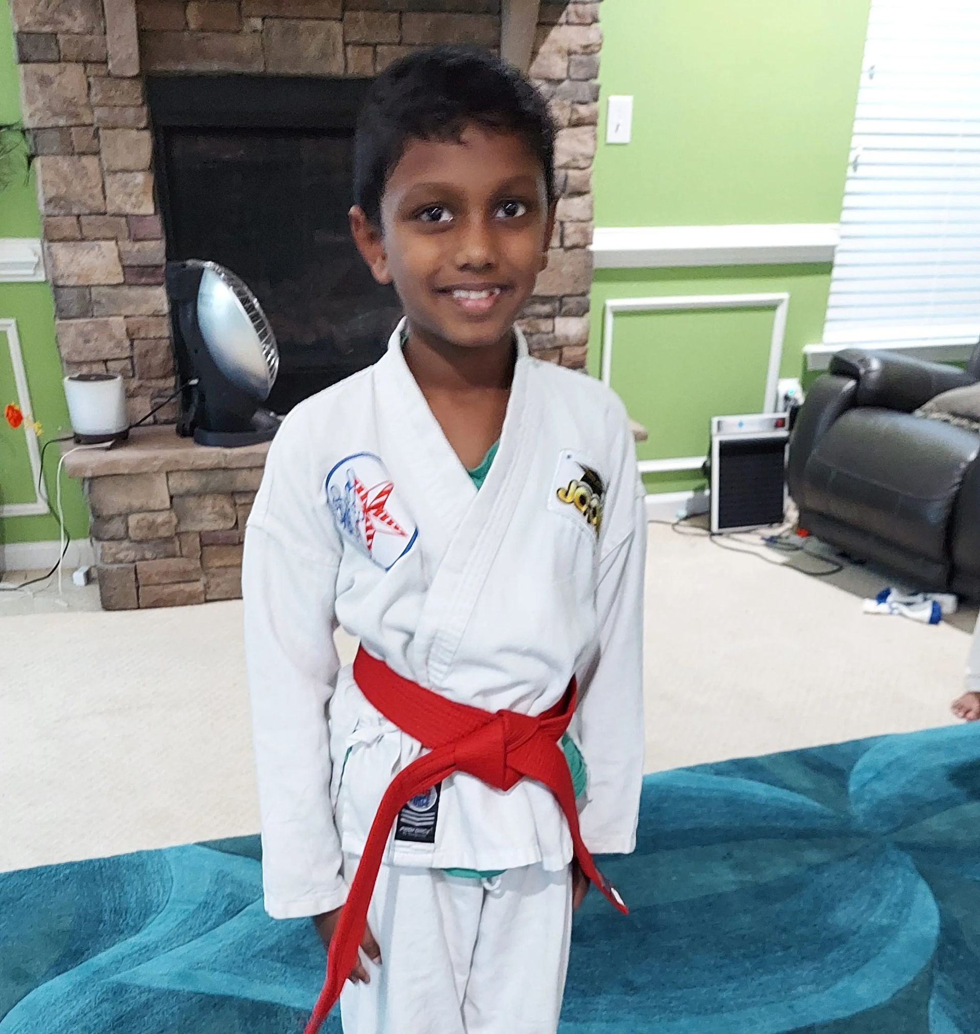 Boy in karate uniform with red belt smiles in front of a fireplace.