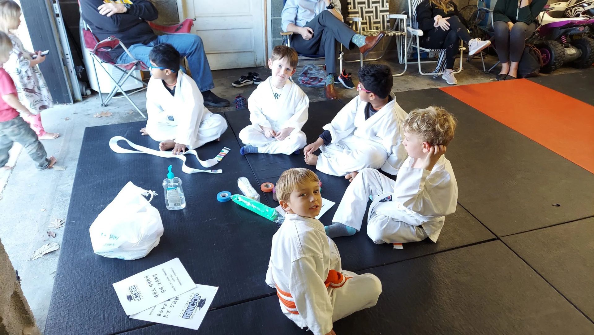 Children in white uniforms sit in a circle on a mat, likely at a martial arts gathering, indoors.