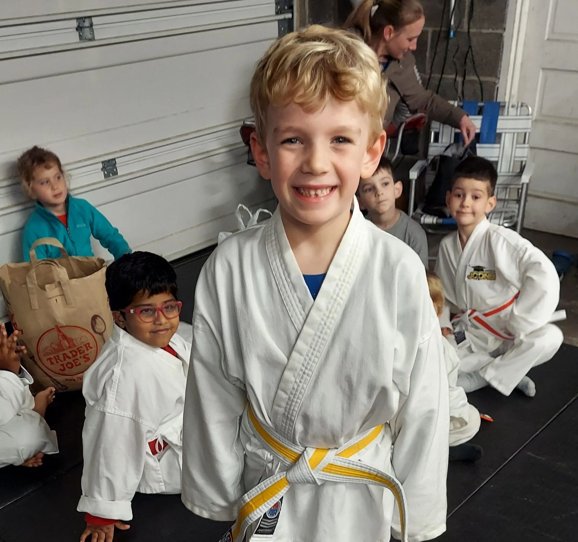 Boy with yellow belt smiles, surrounded by kids in martial arts uniforms. Garage setting.