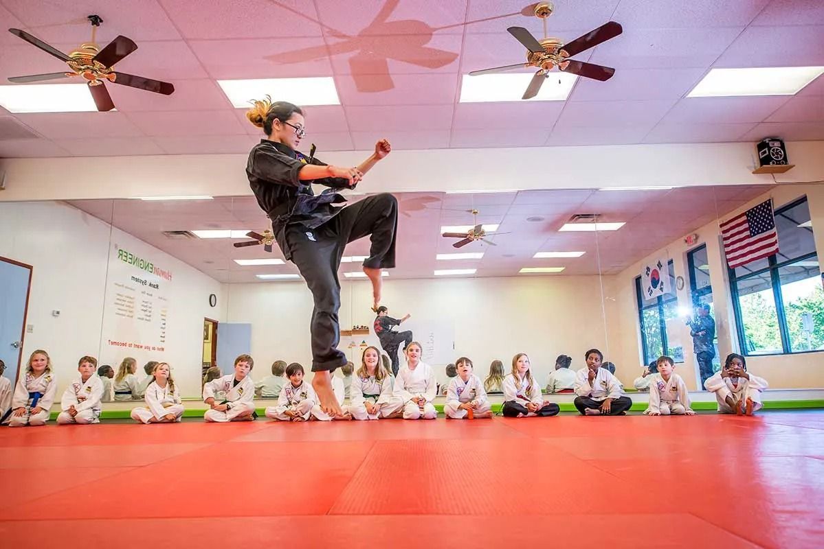 Martial arts instructor in mid-air kick demonstration; students in white uniforms watch. Classroom setting with red mats.