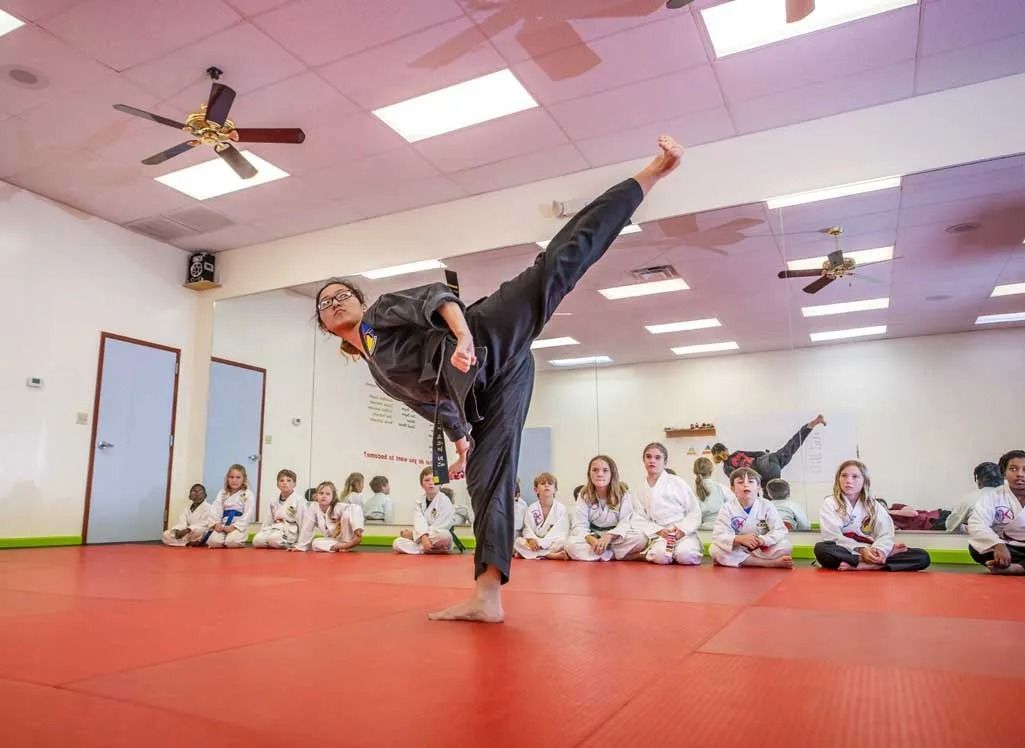 Person in black martial arts uniform performing a high kick on a red mat. Children in white uniforms watch.