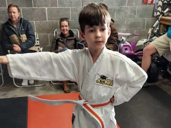 Boy in karate uniform with orange belt, demonstrating a move. People seated in background.