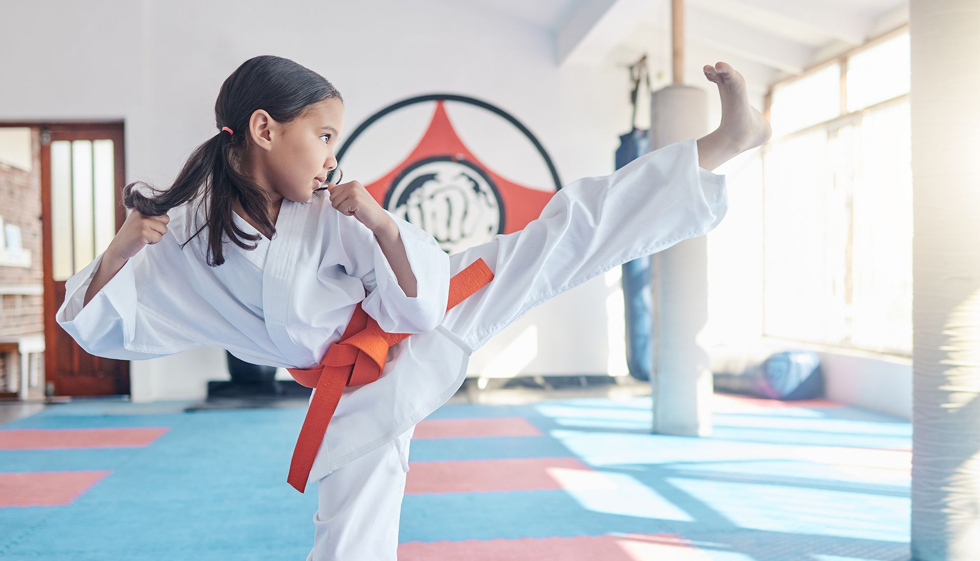 Girl in karate uniform kicking, orange belt, martial arts studio.