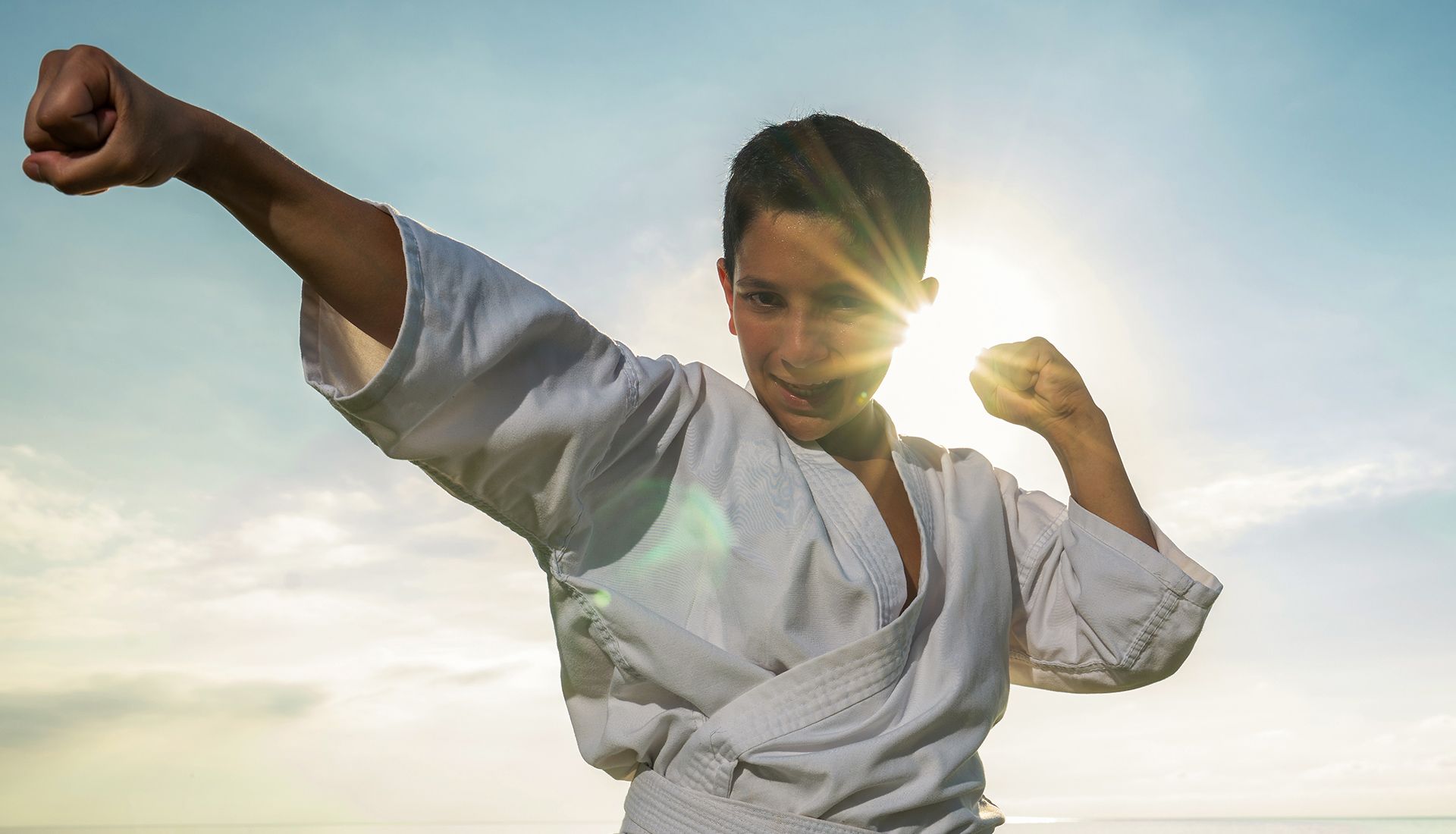 Person in white karate uniform, sunlight behind, throwing a punch with a determined expression.