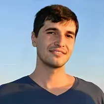 Man with dark hair and a light smile against a blue sky, wearing a dark blue v-neck shirt.