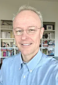 Man with glasses smiling, wearing a blue shirt. Bookshelves are visible in the background.