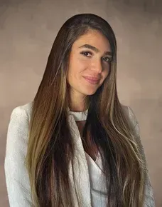 Woman smiling outdoors with brown hair; sunglasses on head; cafe setting.