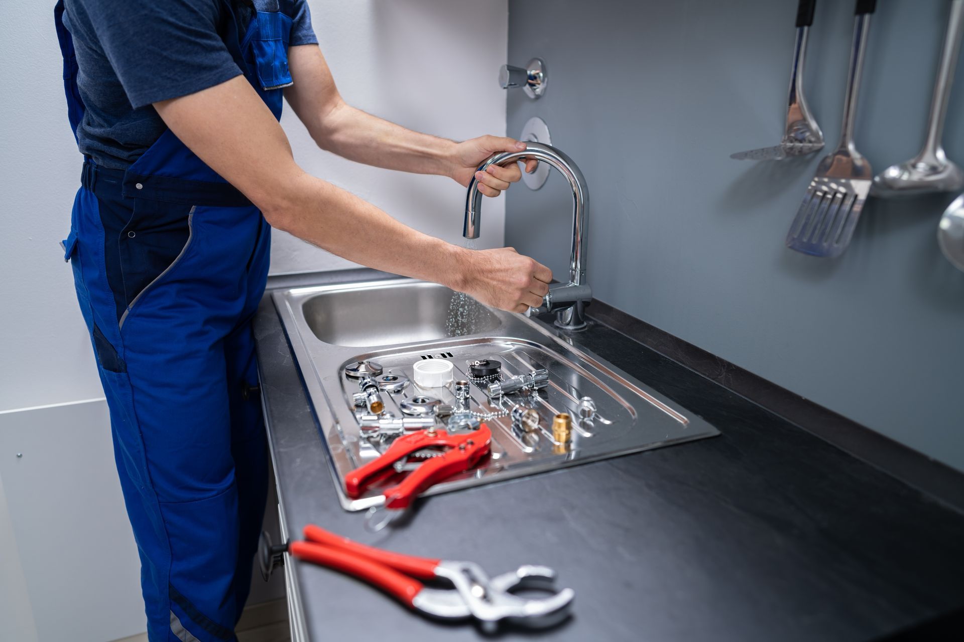 A plumber in blue workwear repairs a kitchen faucet with tools placed in the sink.