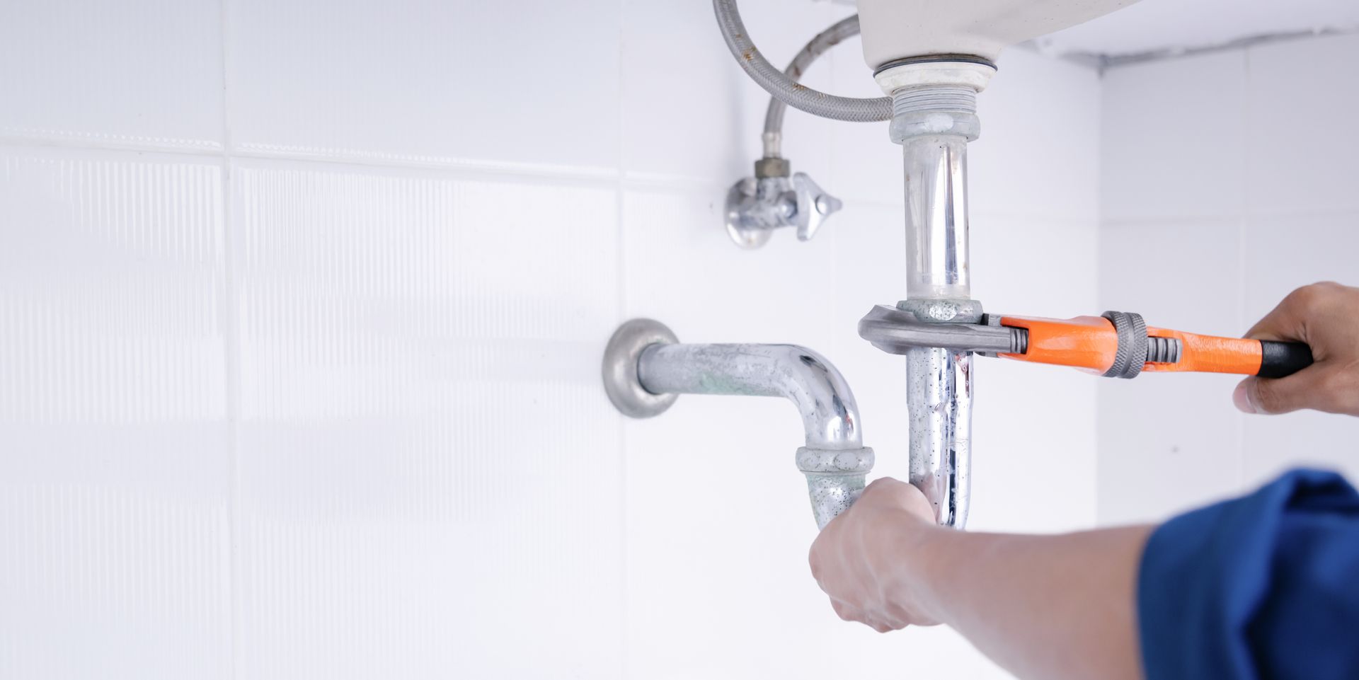 A person uses a wrench to tighten a metal P-trap pipe under a bathroom sink against a white tiled wall.