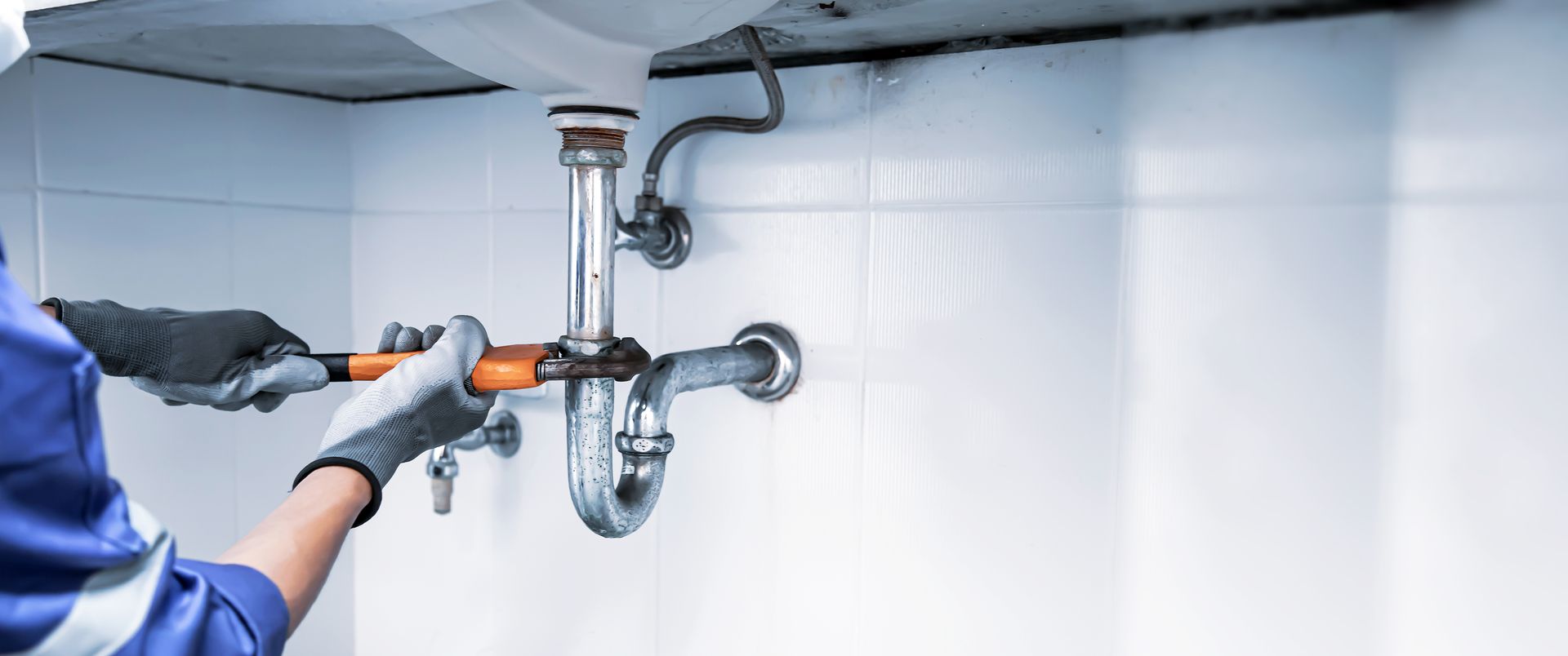A person wearing work gloves uses a wrench to tighten a metal drain pipe under a sink against a tiled white wall.