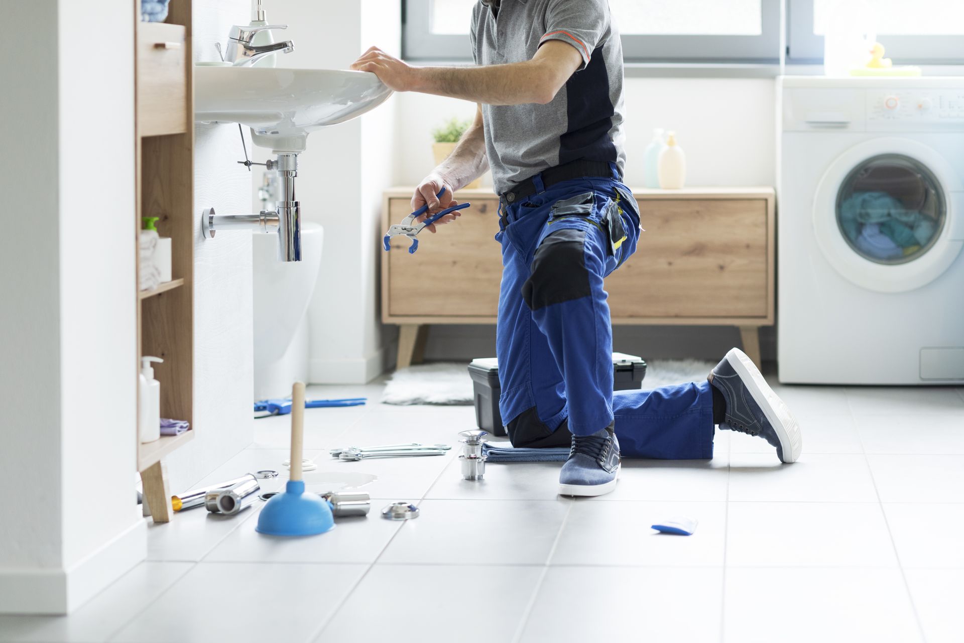 A plumber in blue work pants kneels on a white-tiled floor, repairing a bathroom sink with tools and a plunger nearby. A plumber in blue work pants kneels on a white-tiled floor, repairing a bathroom sink with tools and a plunger nearby.