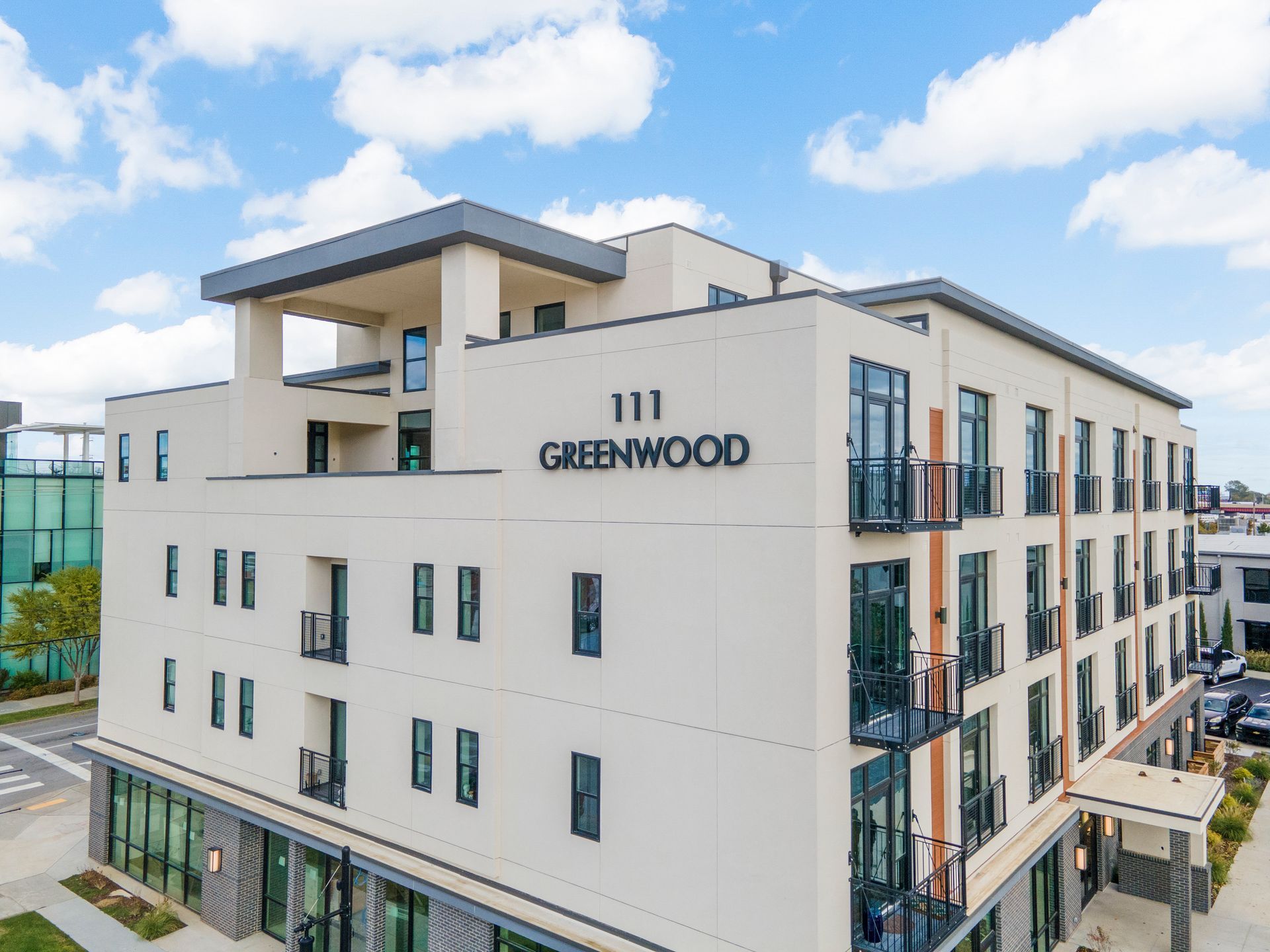 Modern apartment building, 111 Greenwood, with tan exterior, balconies, and street-level retail space on a sunny day.