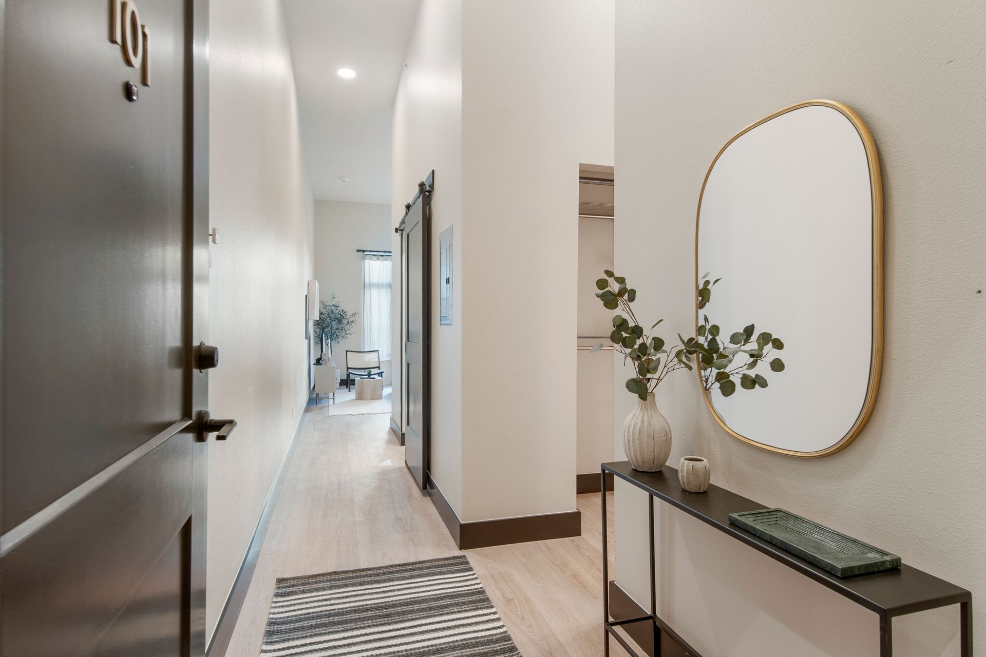 Narrow hallway with light wood flooring, white walls, and a decorative console table with a mirror.