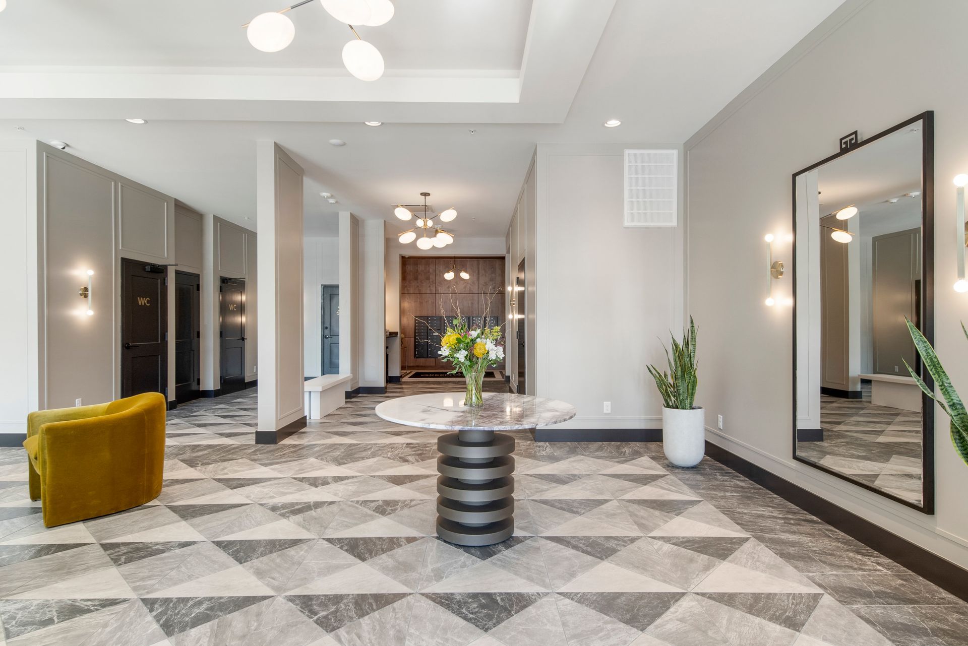 Lobby with geometric tile floor, yellow chair, round table with flowers, large mirror, and multiple light fixtures.