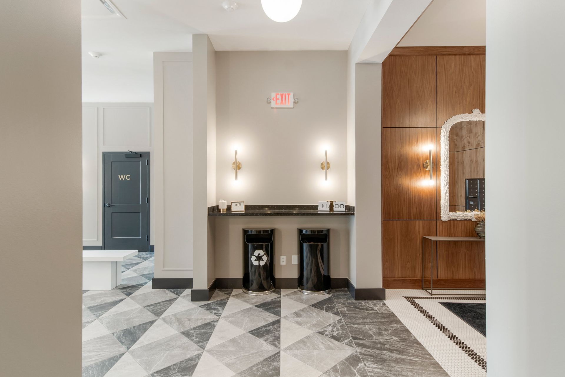 Lobby with exit sign, two wall sconces, two trash bins, geometric tile flooring, wood paneling, and a mirror.