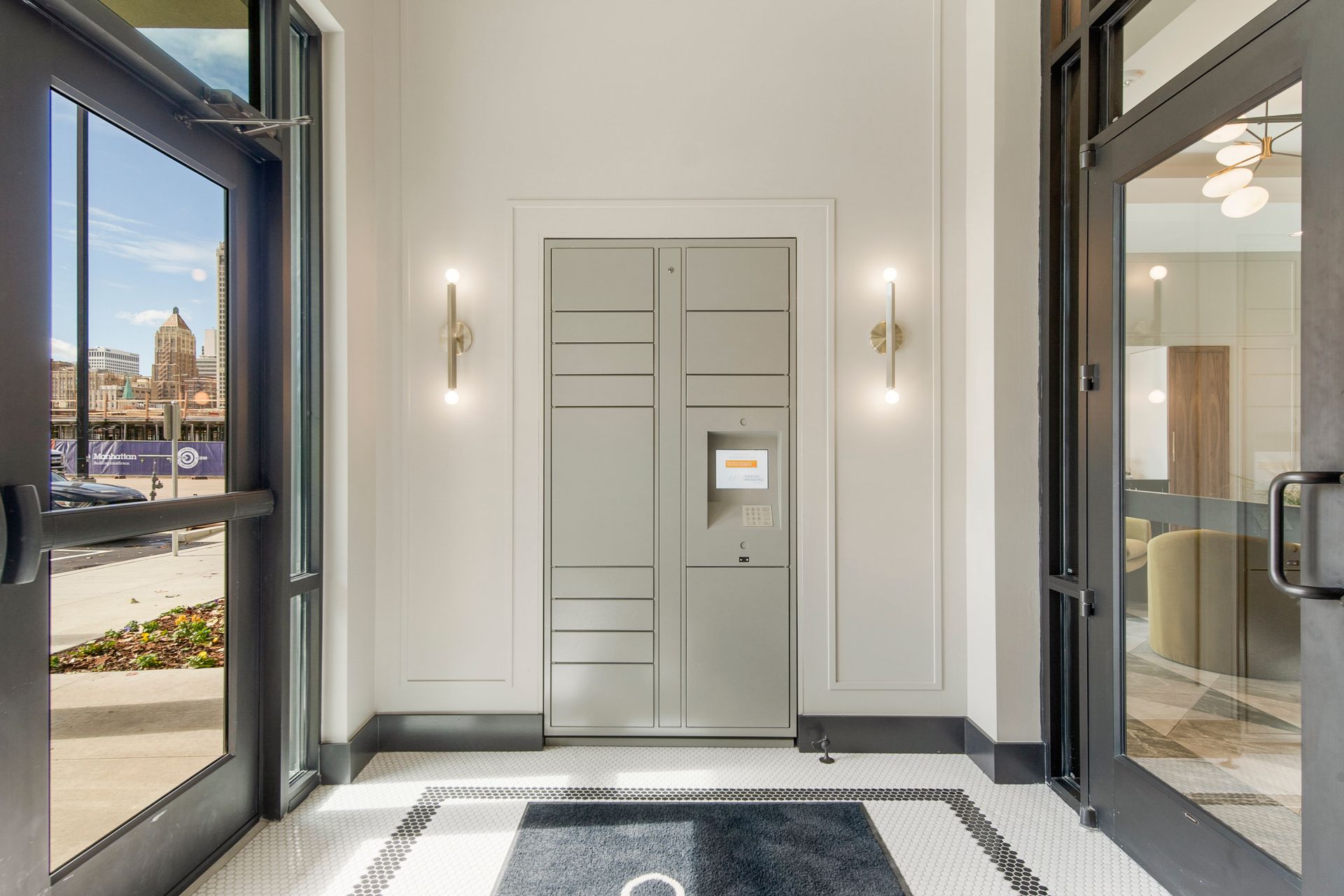 Gray package lockers in a well-lit lobby, flanked by glass doors, sconces, and a patterned floor.