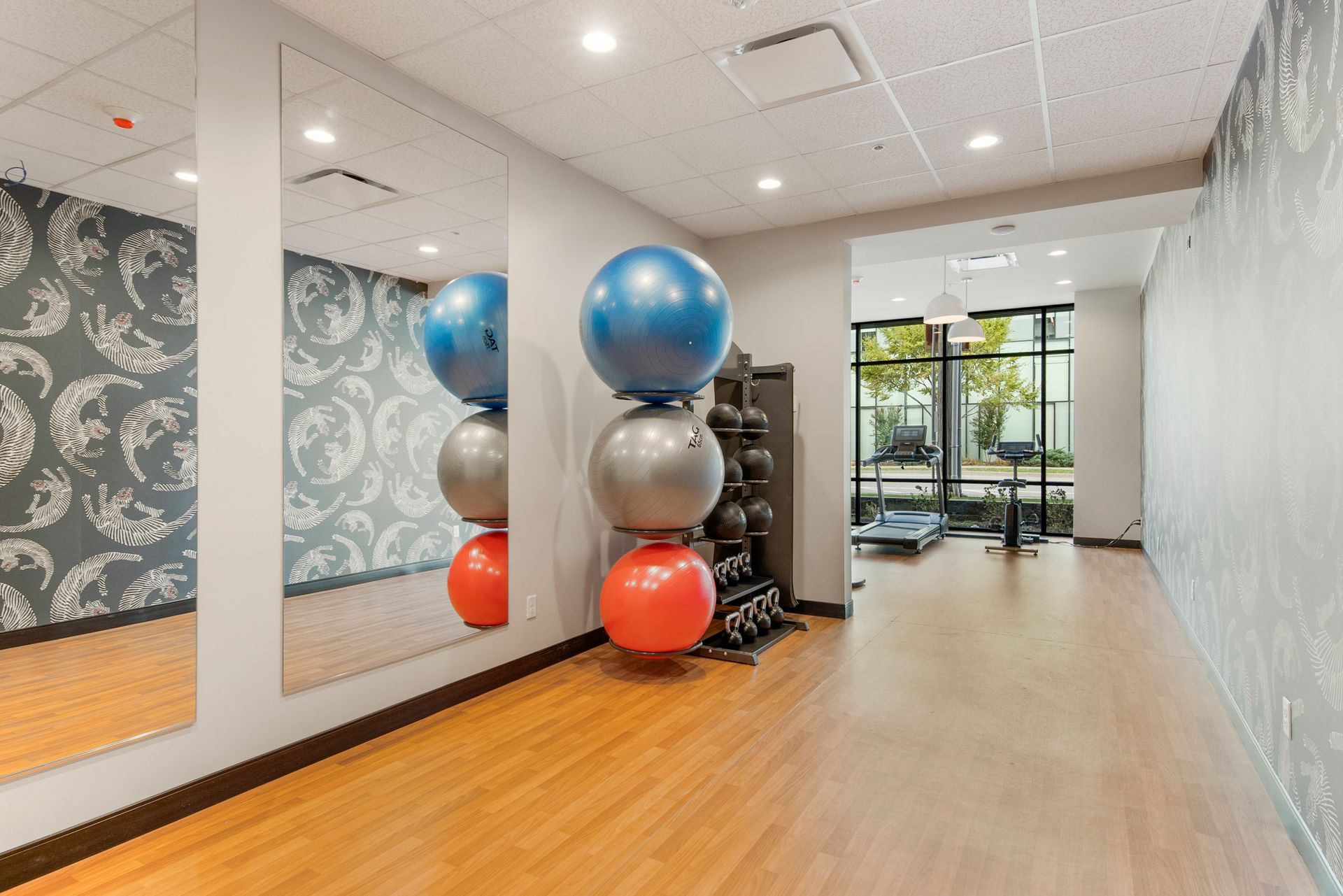 Gym with exercise balls stacked, dumbbell rack, mirrors, wood floor, patterned wall, and a view to outside.