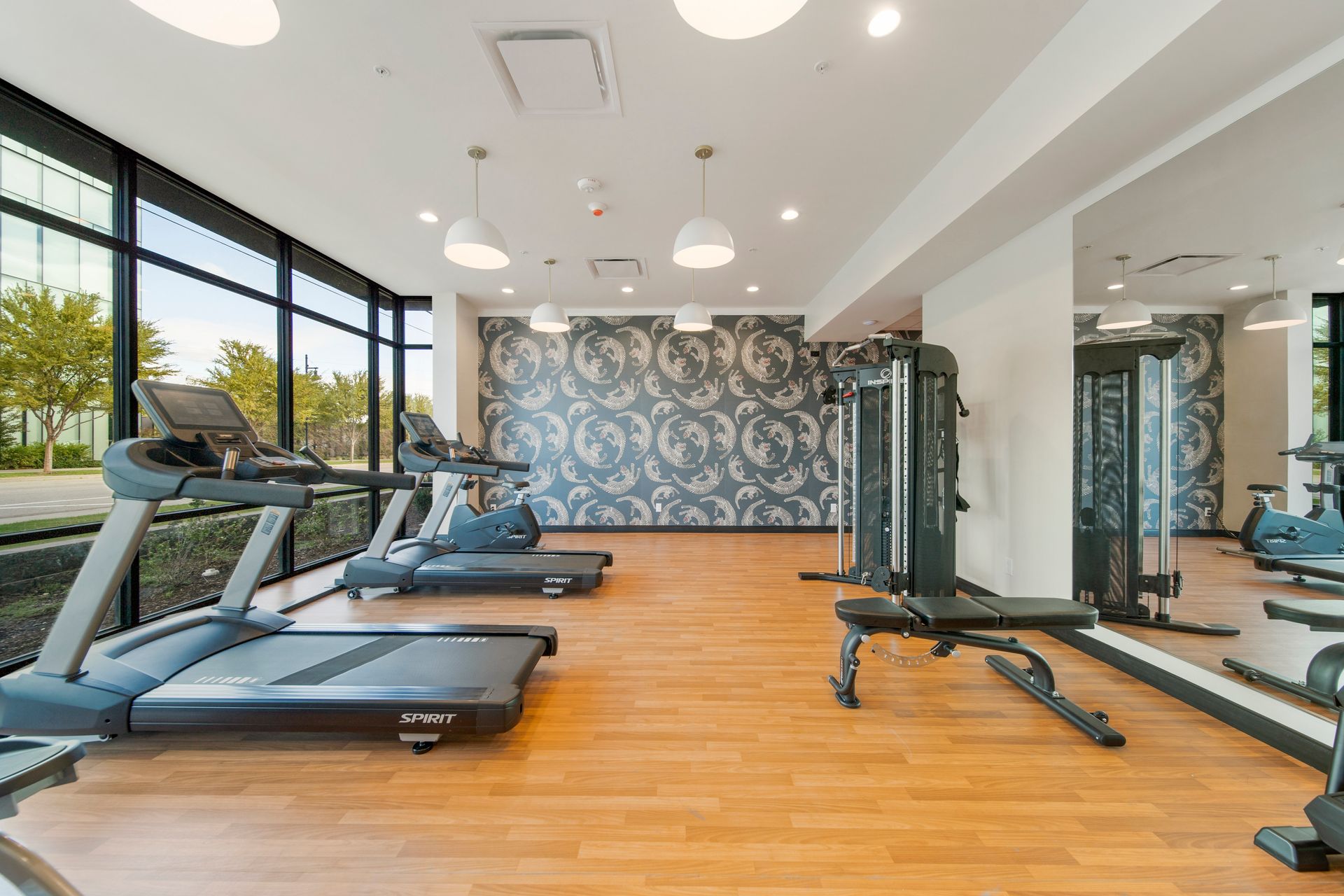 Gym with treadmills, weight machines, and large windows. Wooden floor, patterned wall, and overhead lights.