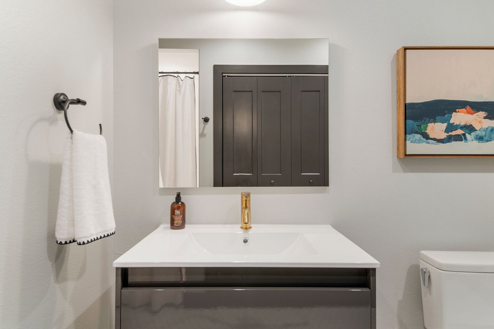 Bathroom with gray vanity, gold faucet, square mirror, towel, and abstract artwork.
