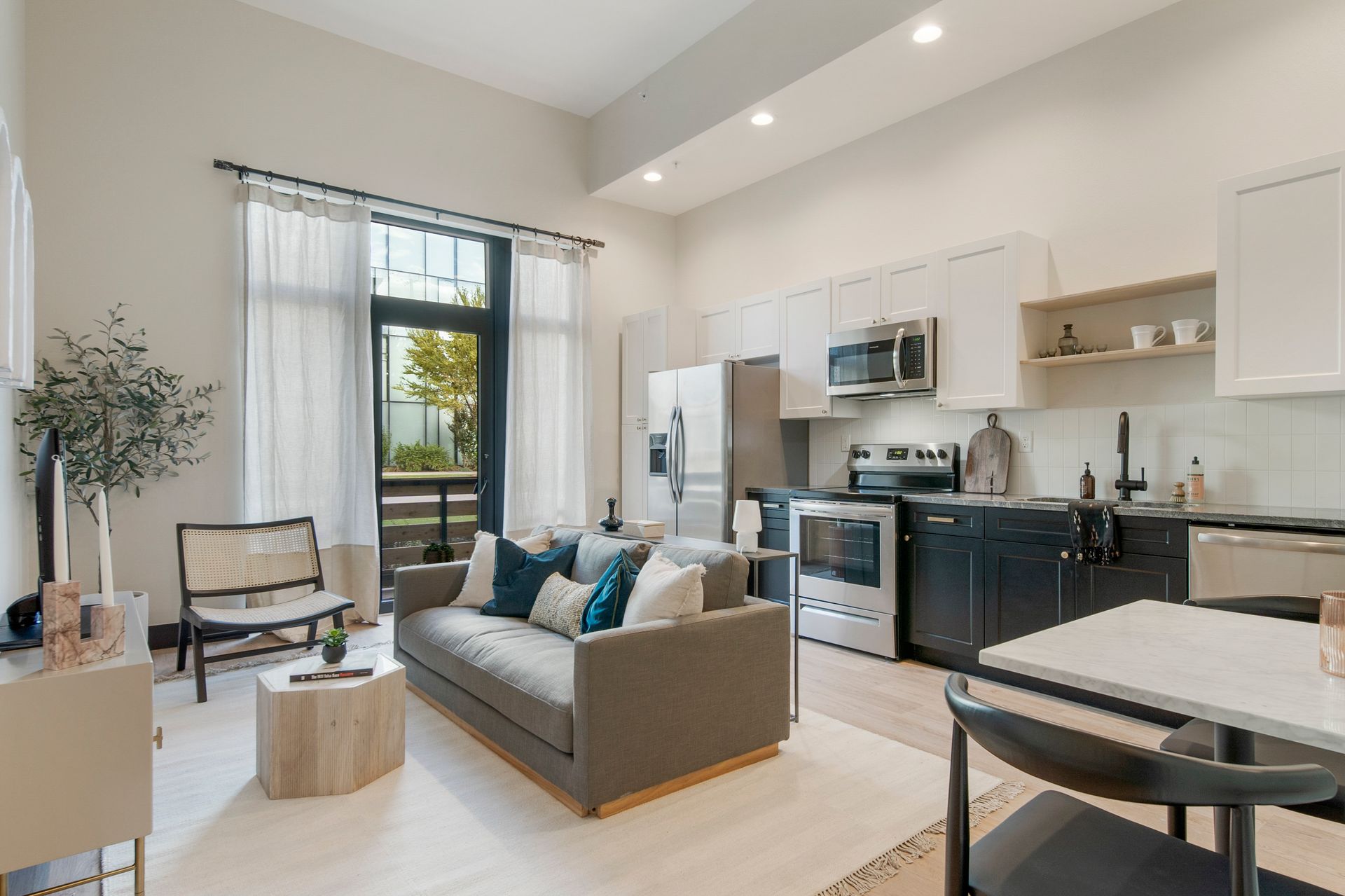 Cozy living space with kitchen; gray sofa, white walls, large window with sheer curtains.