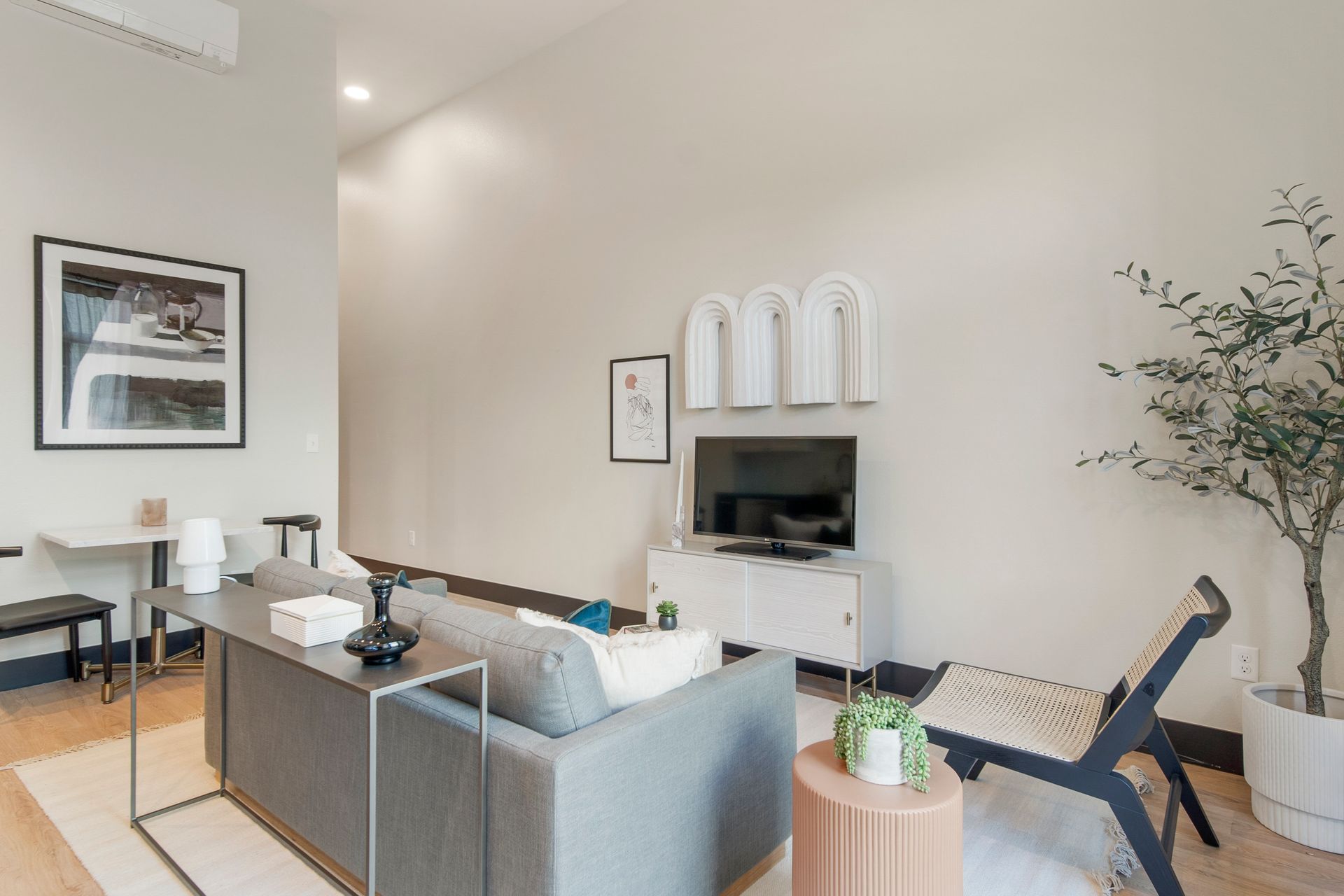 Modern living room with a gray sofa, TV, white walls, and a small dining area.