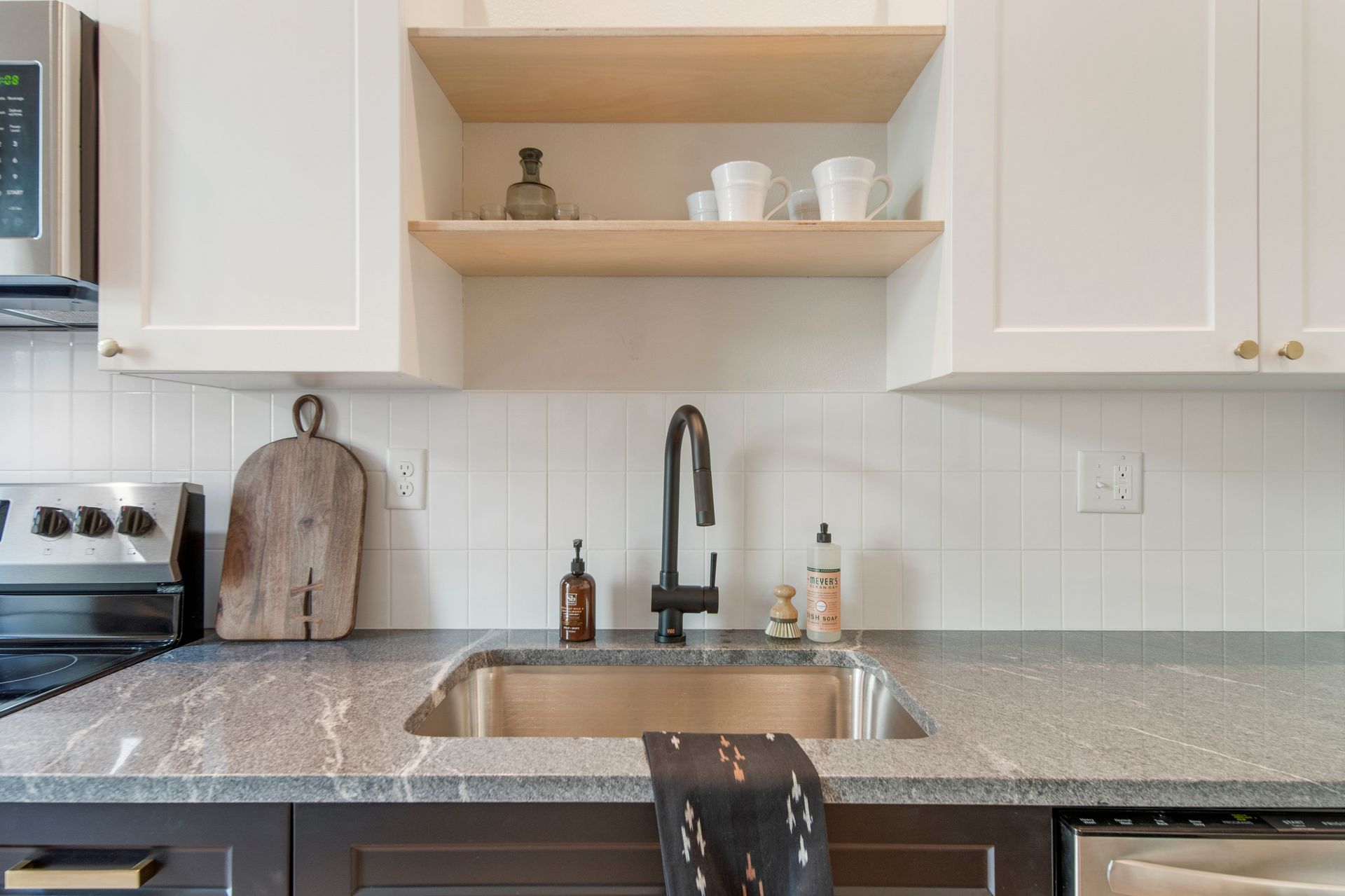 Kitchen with white cabinets, wood shelves, dark faucet, gray countertop, and stainless steel sink.