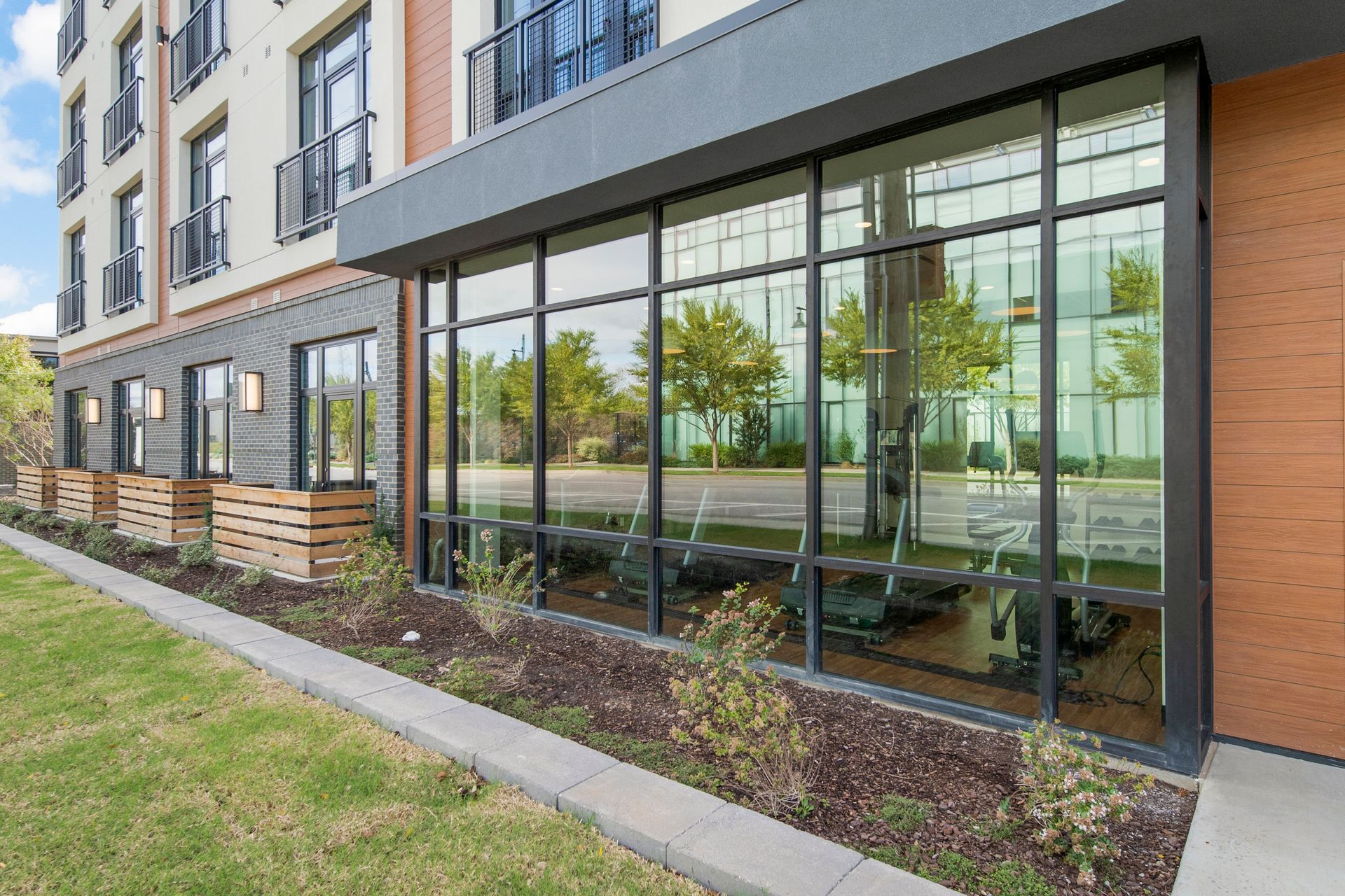 Building exterior with large windows reflecting trees, brick and gray facade, and grassy area.