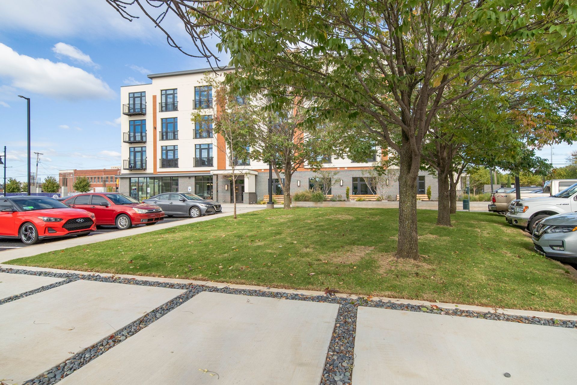 Modern apartment building with cars parked in front, green grass, and trees. Blue sky.