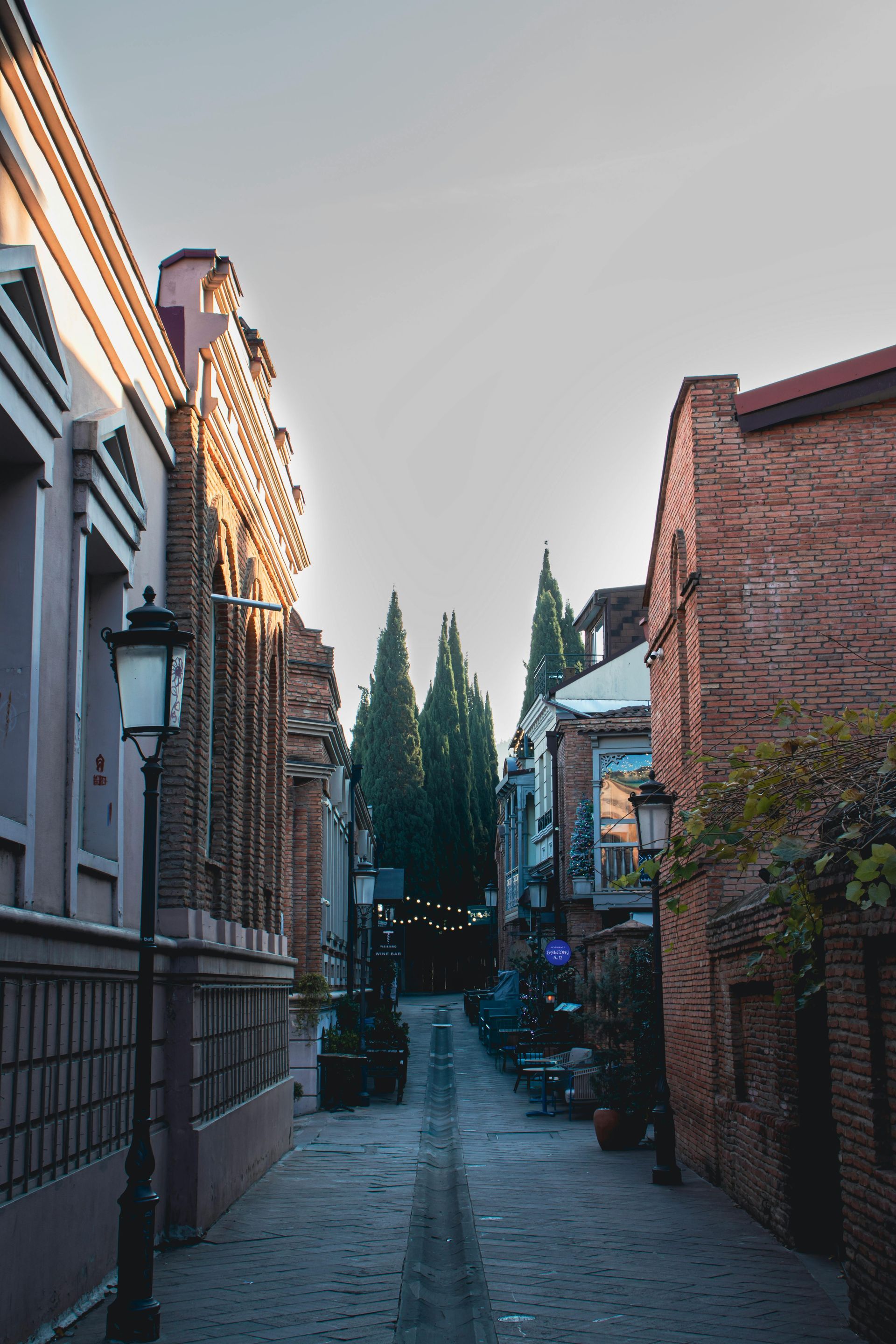Cobblestone alleyway lined with buildings, street lamps, and trees in the distance; warm sunlight.