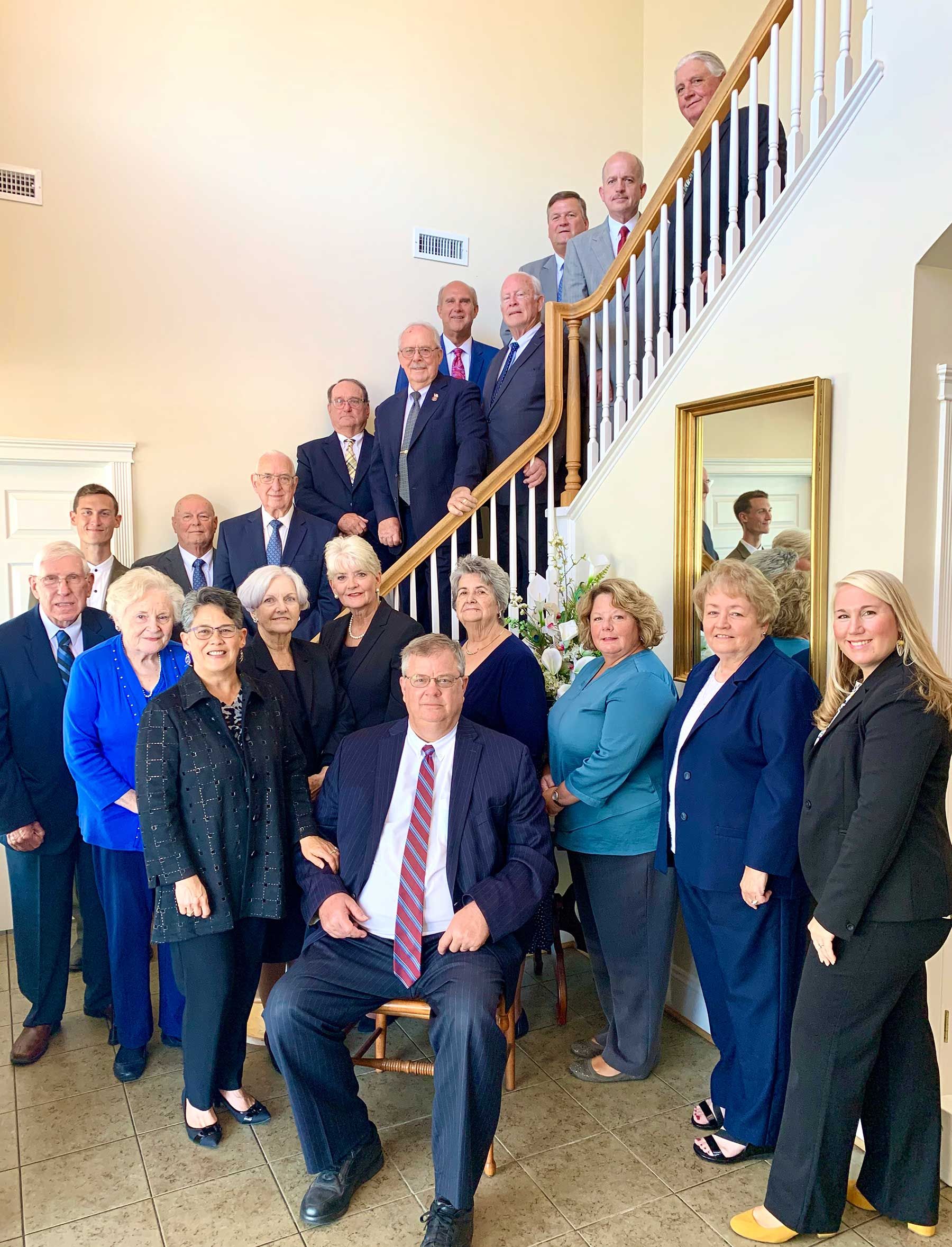 Group of people pose on a staircase, mostly in suits and dresses, smiling for a photo indoors.