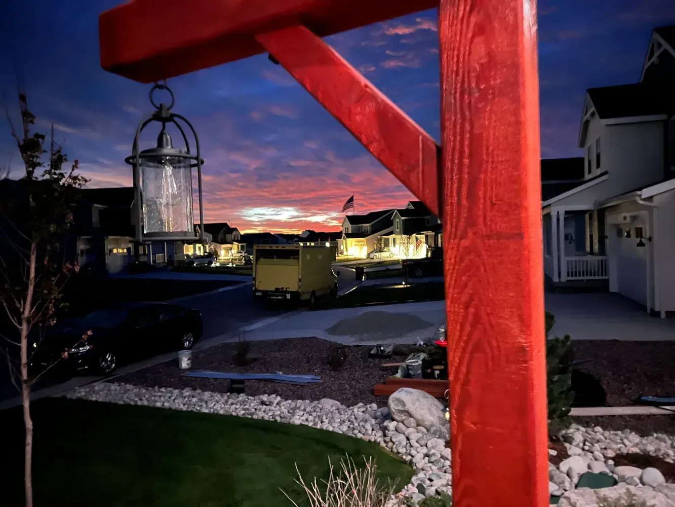 Red wooden structure holding a lantern. Neighborhood street scene at dusk with colorful sky.