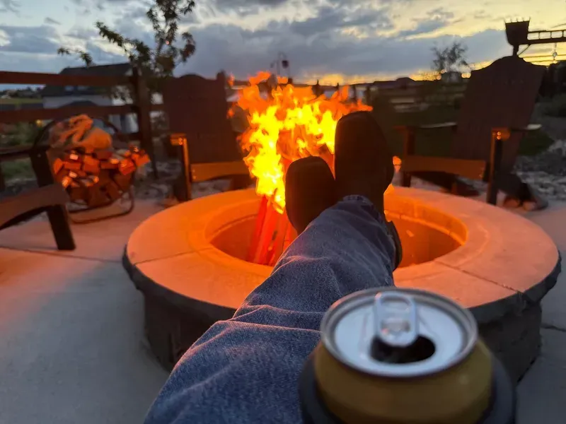 Person relaxing by a fire pit, holding a can. Sunset in the background.
